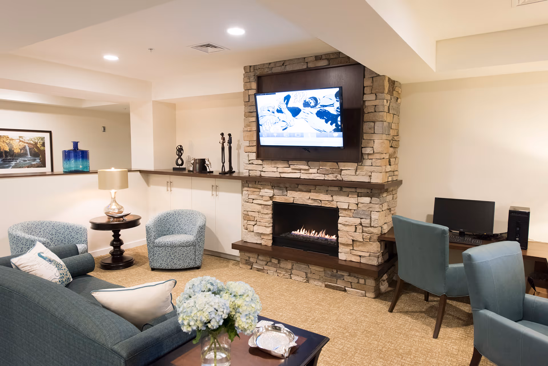 A cozy living room area with a stone fireplace and a mounted flat-screen TV above it. The room features a blue sofa with decorative pillows, two blue patterned armchairs, a small round wooden side table with a lamp, and a coffee table with a vase of blue and white flowers. There is also a desk with a computer and two blue chairs. The walls are light-colored, and there is a framed picture and decorative items on a shelf behind the seating area.