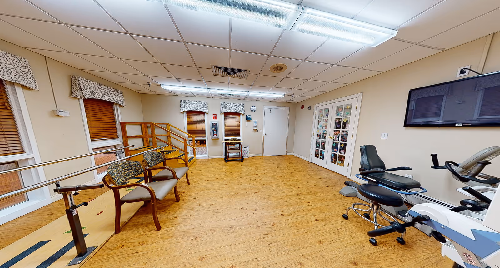 Spacious rehab/exercise room with wooden floors, parallel walking bars, seating, exercise bikes, and a wall-mounted TV.