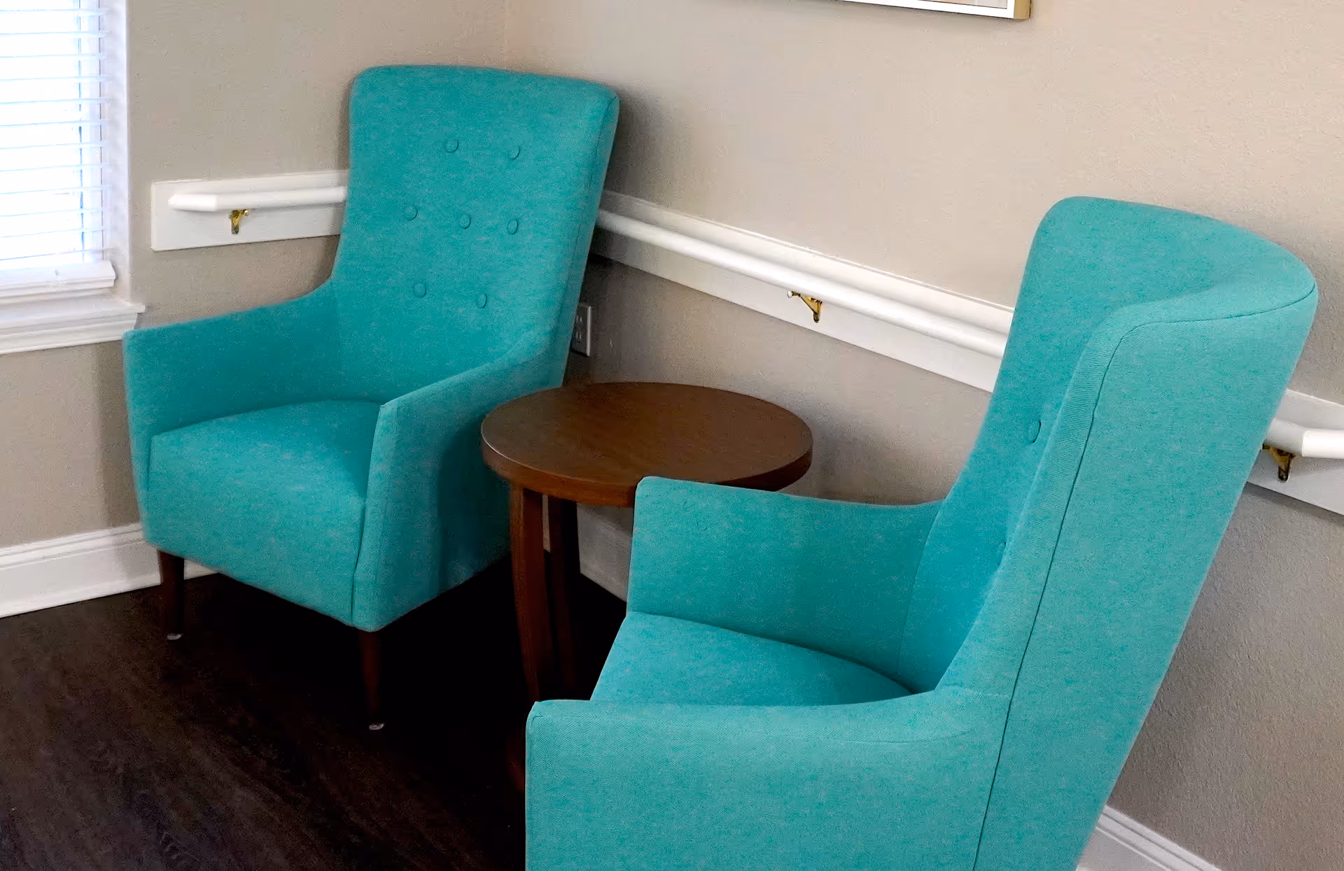 Two teal upholstered armchairs with button tufting on the backrests are positioned around a small round wooden side table in a corner of a room with beige walls and dark wood flooring. A window with white blinds is visible on the left side.