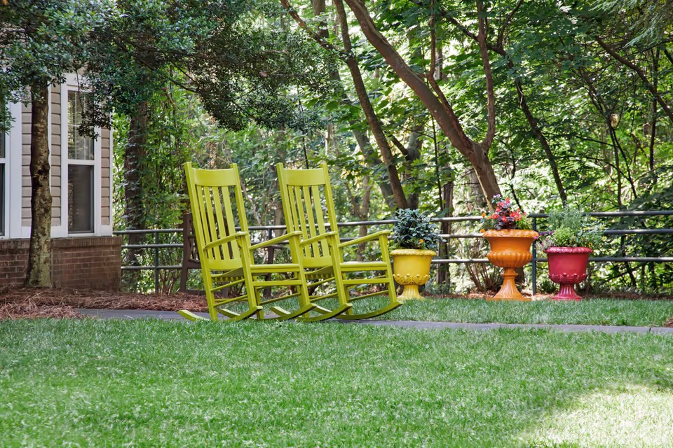 Two lime-green rocking chairs sit on a lawn beside colorful potted plants and trees near a building.