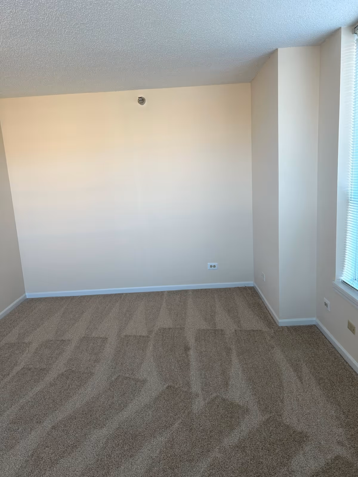 Empty room with beige walls and carpeted floor with vacuum lines, a window with blinds on the right side, and electrical outlets on the walls.