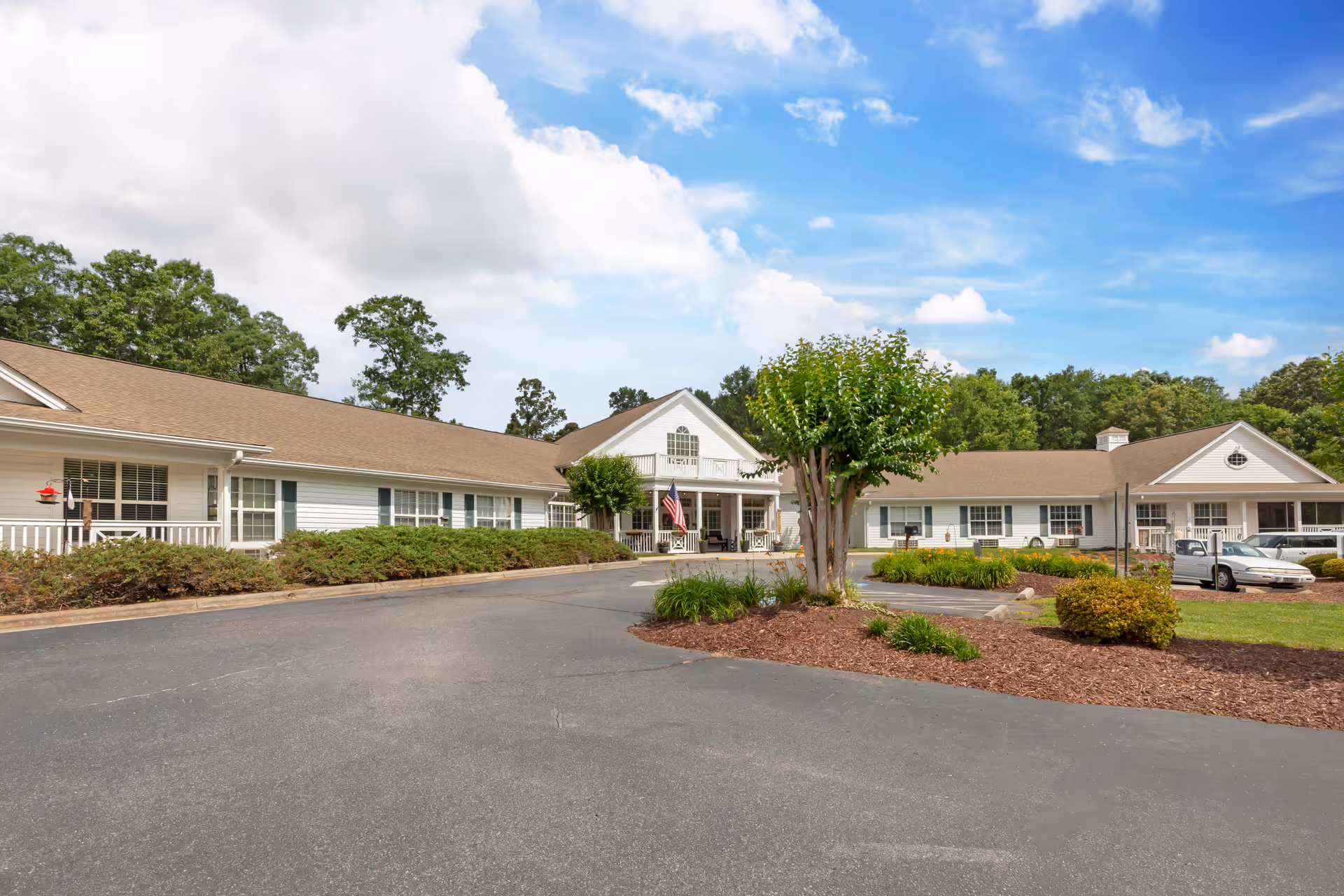 Exterior view of a single-story senior living facility with white siding and a brown roof. The building is surrounded by landscaped bushes, small trees, and a paved driveway. The sky is partly cloudy with blue patches visible.