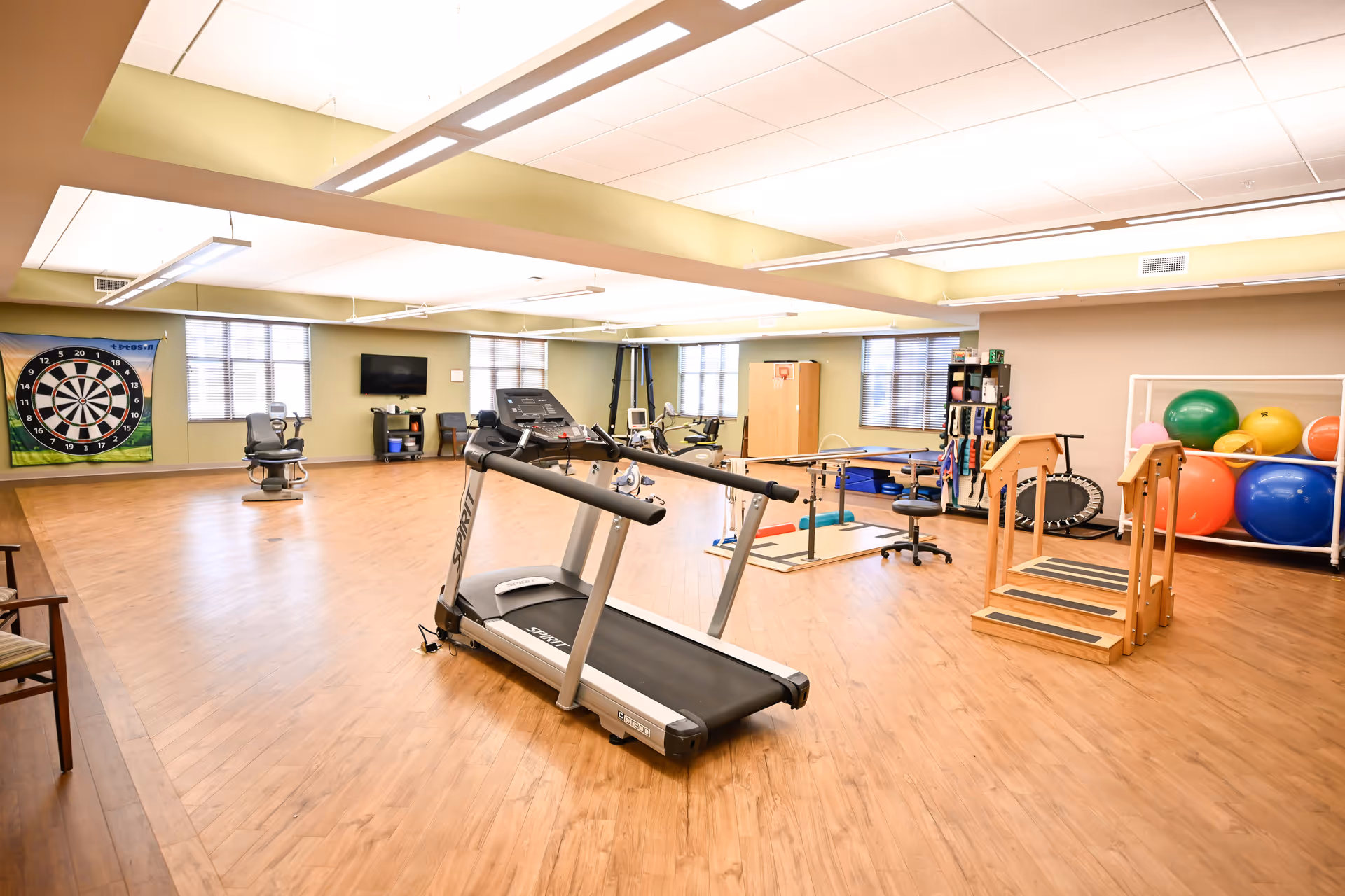 Bright senior living exercise/therapy room with a treadmill, therapy equipment, stability balls and a dartboard.