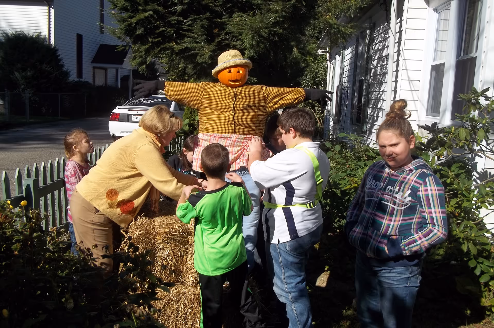 A group of children and an adult woman are gathered outside near a white house, decorating a scarecrow dressed in a yellow checkered shirt and a straw hat. The scene is sunny with greenery and a white picket fence in the background.