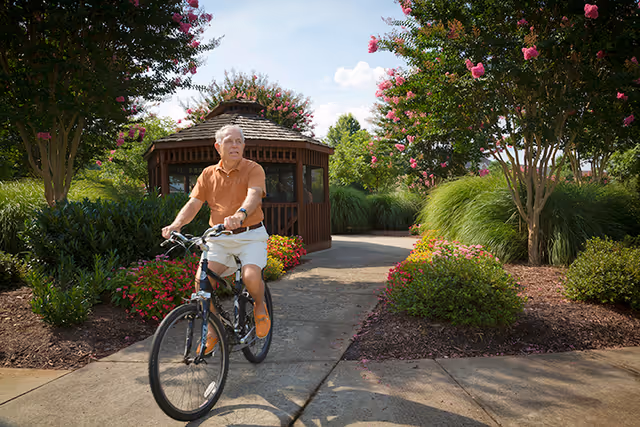 An elderly man riding a bicycle on a paved path in a garden area with flowering bushes and trees, with a wooden gazebo in the background under a partly cloudy sky.