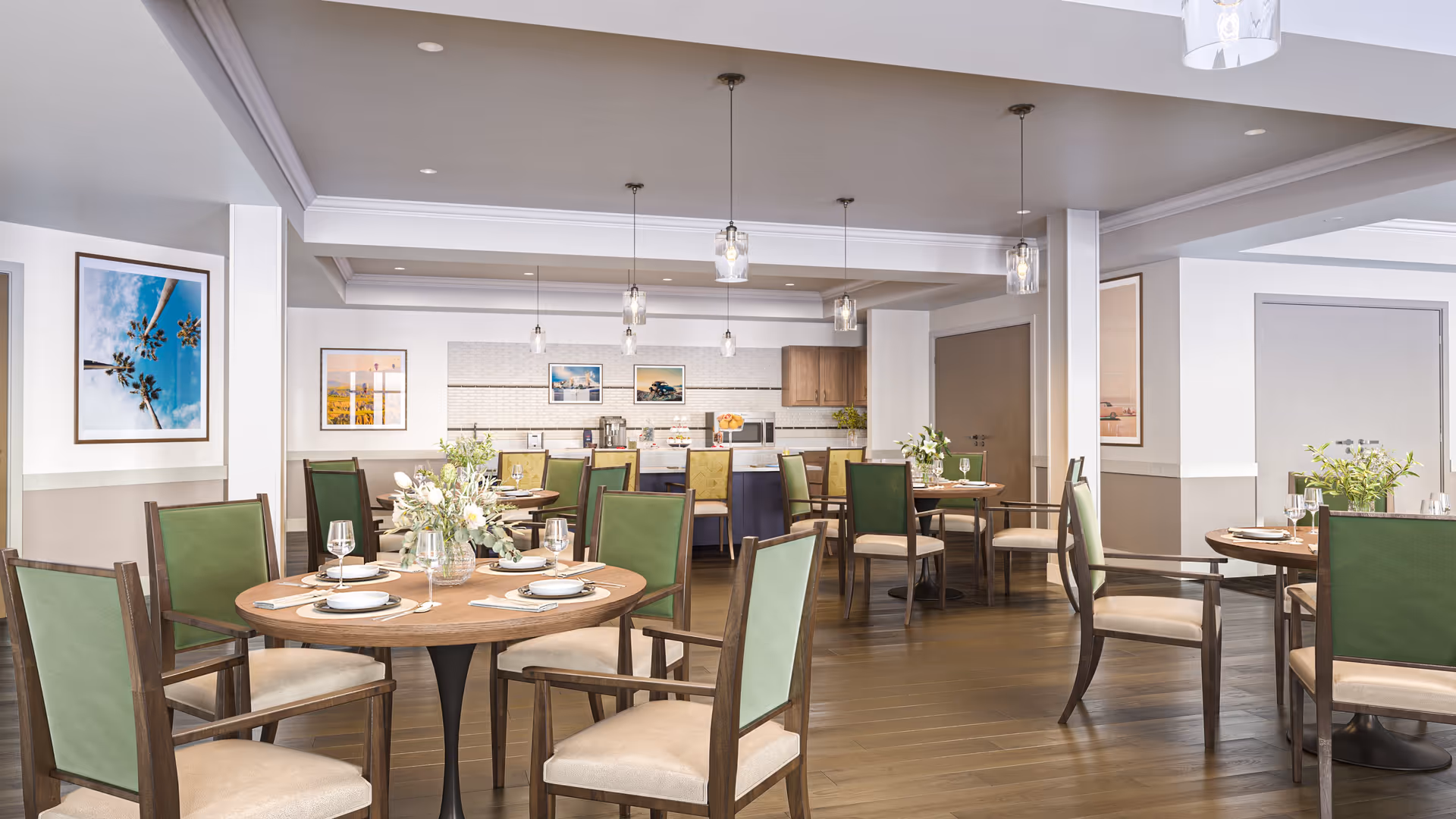 A bright and modern dining room with round wooden tables set with plates, glasses, and cutlery. The chairs have green backs and beige seats. The room features pendant lights hanging from the ceiling, framed artwork on the walls, and a kitchen area in the background with cabinets, a microwave, and a coffee machine.
