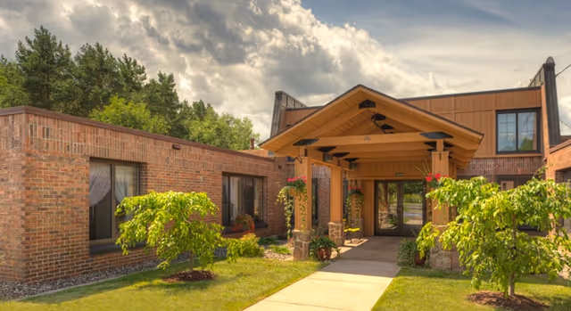 Front entrance of a brick senior living facility with a covered wooden porte-cochère, walkway, and landscaped greenery under a partly cloudy sky.