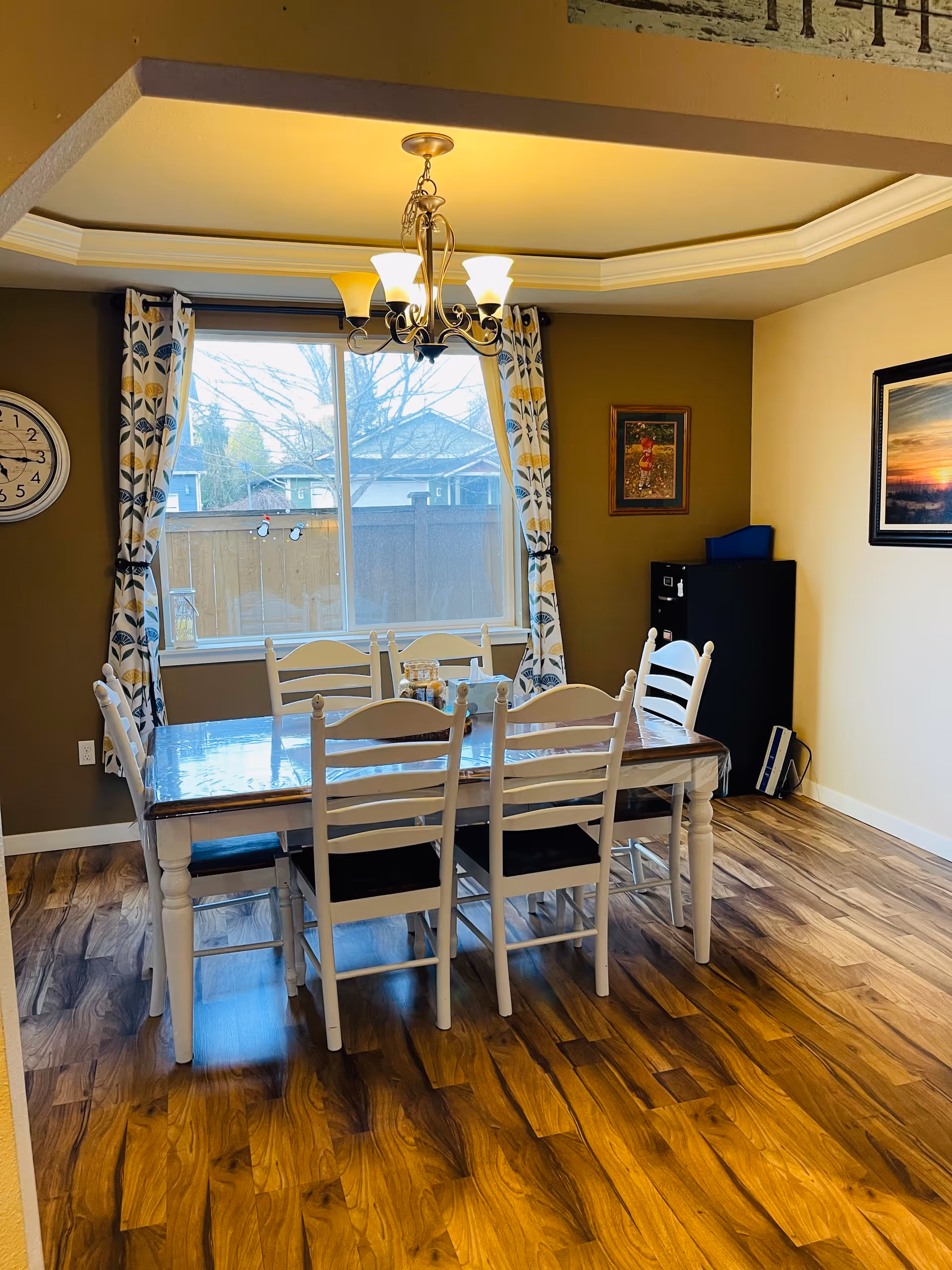 Bright dining room with a wooden table and six white chairs under a chandelier by a large window.