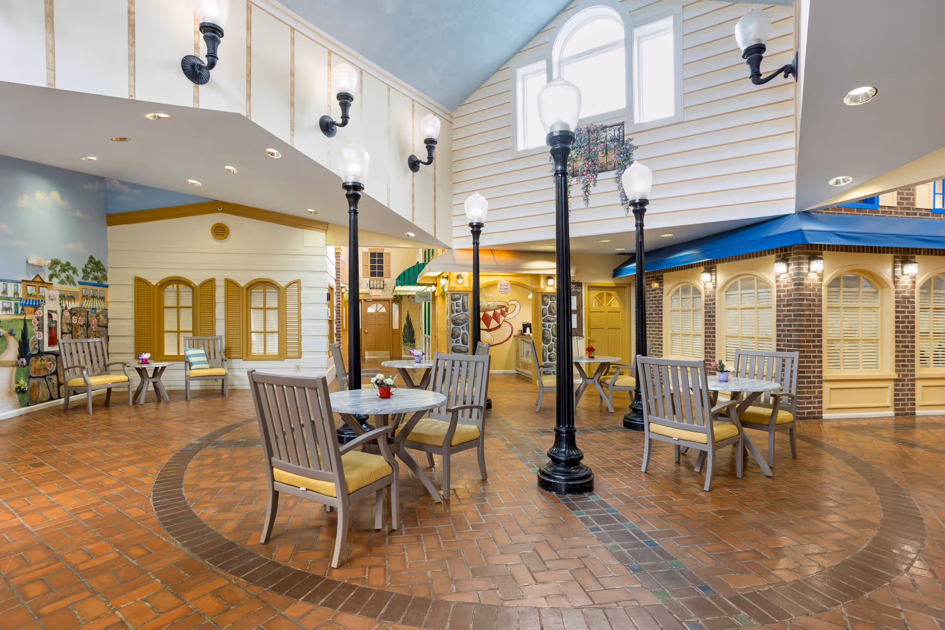 Indoor seating area designed to resemble an outdoor village street with brick flooring, street lamps, tables with chairs, and building facades with windows and shutters. The space is well-lit with natural light from large windows above and wall-mounted lamps.