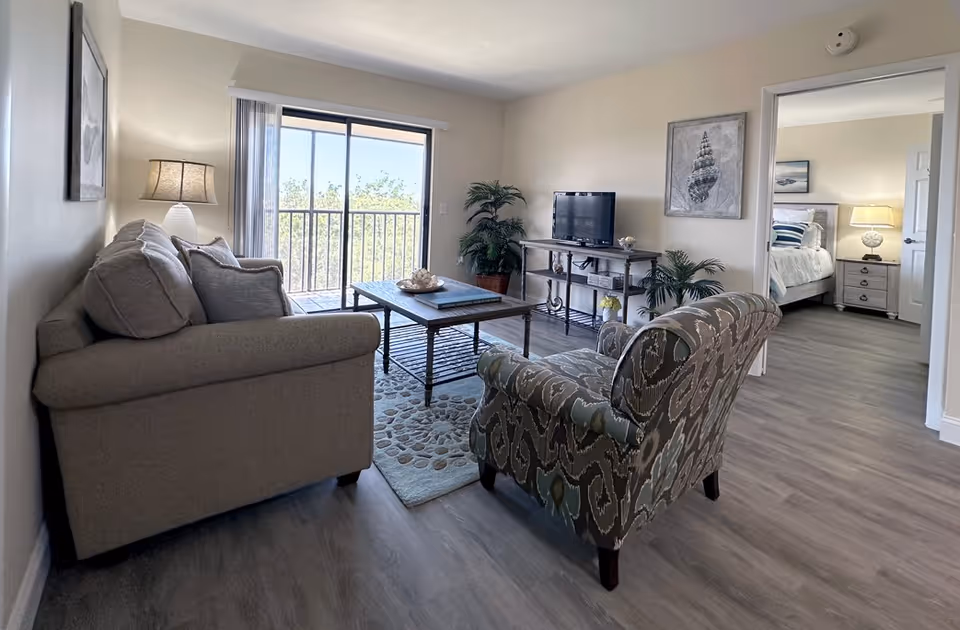 Bright living room with sofa, patterned chair, coffee table, sliding balcony door, and TV.
