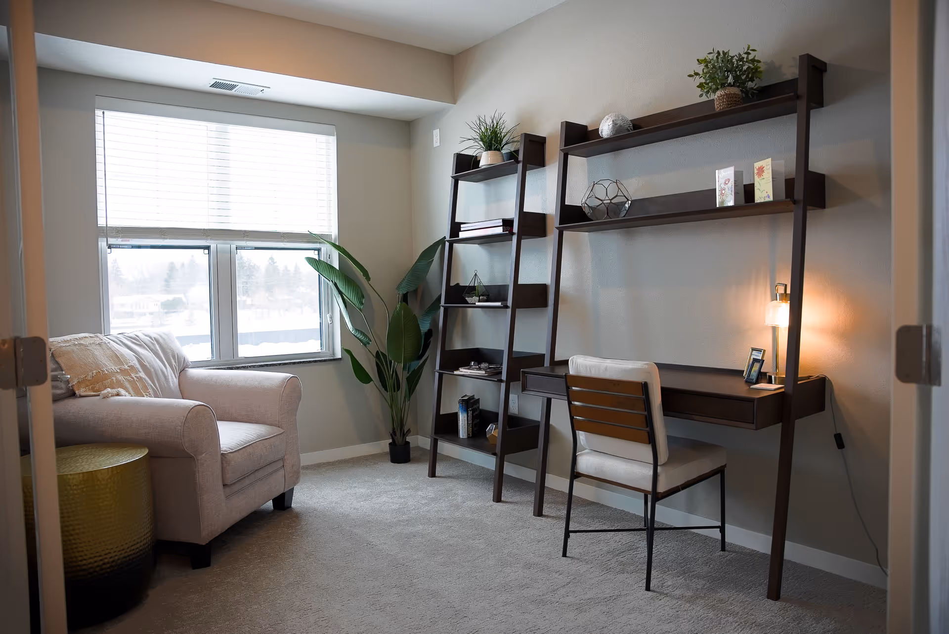 A cozy room with a beige armchair next to a window with white blinds, a tall green plant in the corner, and a dark wooden desk with a matching chair. The desk has a small lamp and decorative items on the shelves above it. The room has light-colored walls and carpeted flooring.