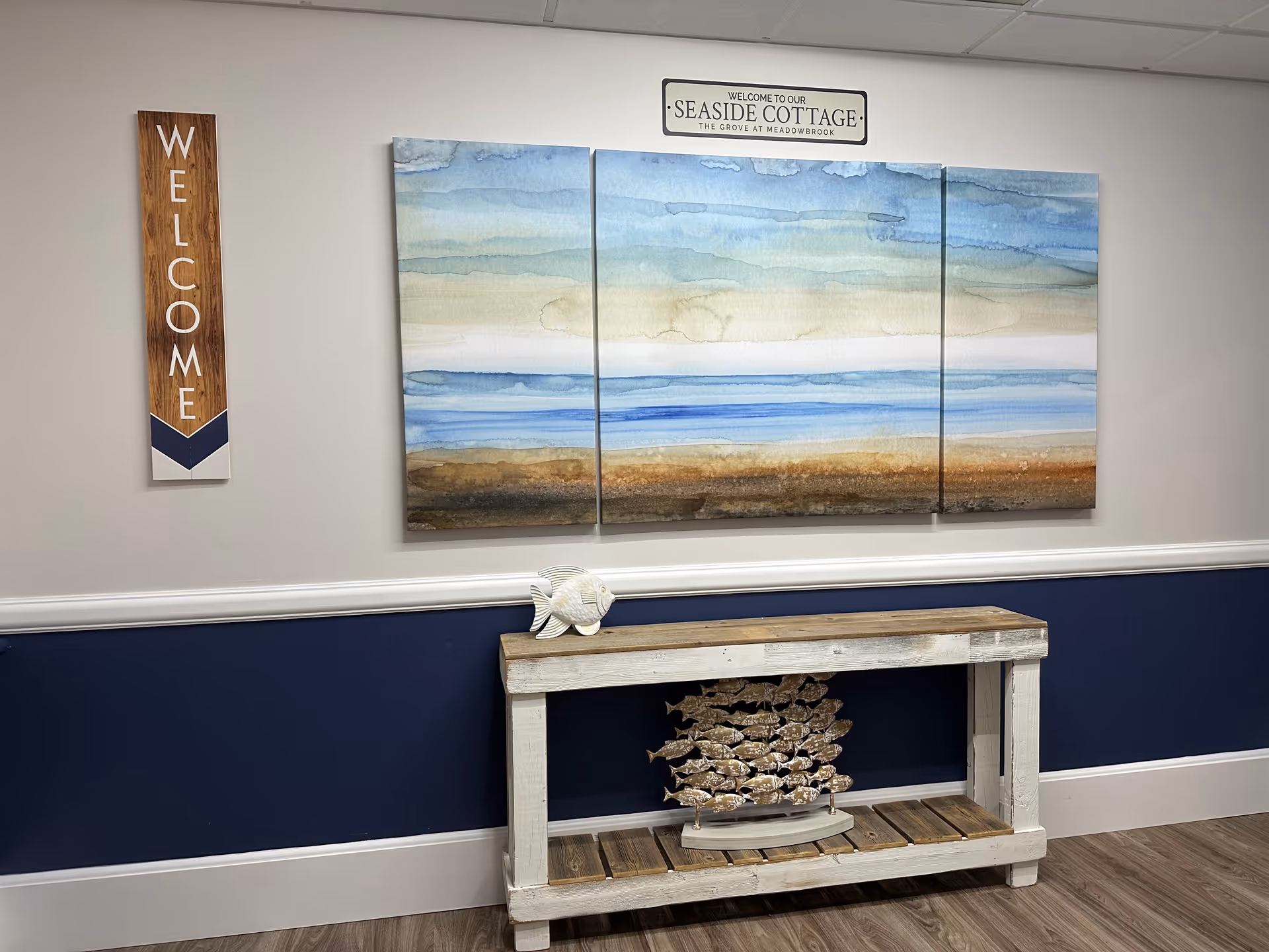 Nautical-themed interior hallway with a three-panel seaside painting, a vertical "WELCOME" sign, and a rustic console table topped with decorative fish.