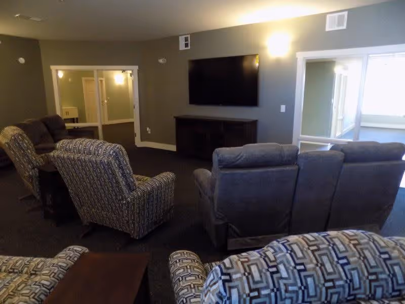 A cozy living room area with multiple upholstered armchairs and recliners arranged facing a wall-mounted flat screen TV above a wooden media console. The walls are painted gray and there are large windows and glass doors letting in natural light from an adjacent room.