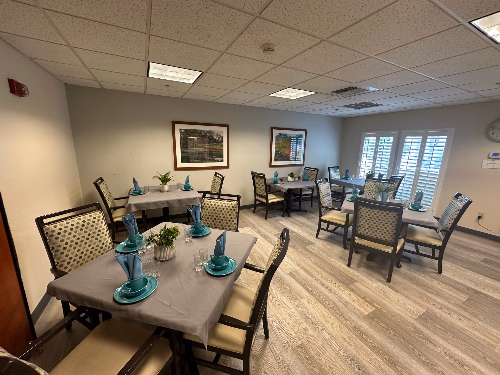 A dining room with several tables covered with gray tablecloths, each set with turquoise plates, glasses, and blue folded napkins. The chairs have patterned upholstery. The room has light wood flooring, white walls with two framed landscape paintings, and windows with white shutters letting in natural light.