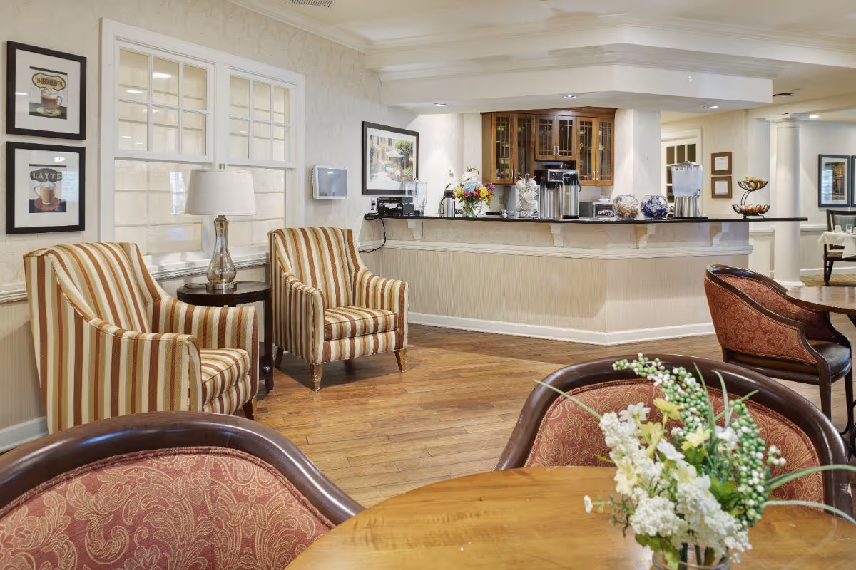 Bright communal dining/lounge area with striped armchairs, round wooden tables with a flower centerpiece, and a service counter in the background.