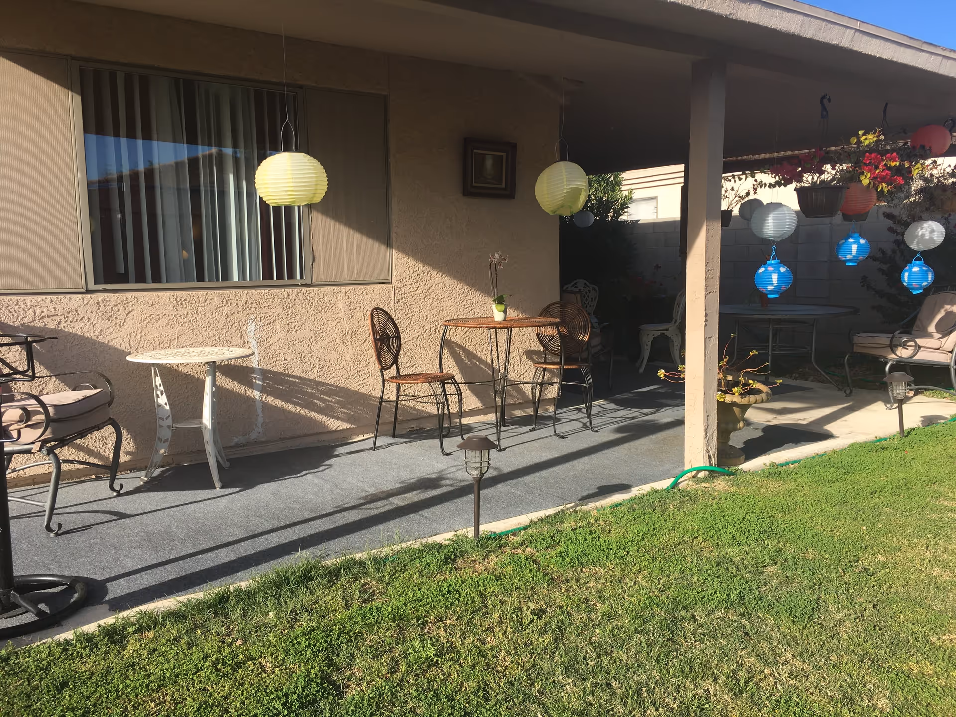 Outdoor patio area with a covered porch featuring several metal chairs and tables. Hanging paper lanterns in yellow, blue, and white colors decorate the space. There are potted plants and flowers hanging from the porch roof, and a grassy lawn in the foreground.