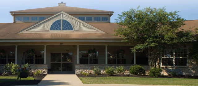 Front exterior view of a single-story building with a peaked roof and large windows, surrounded by greenery and a paved walkway leading to the entrance.