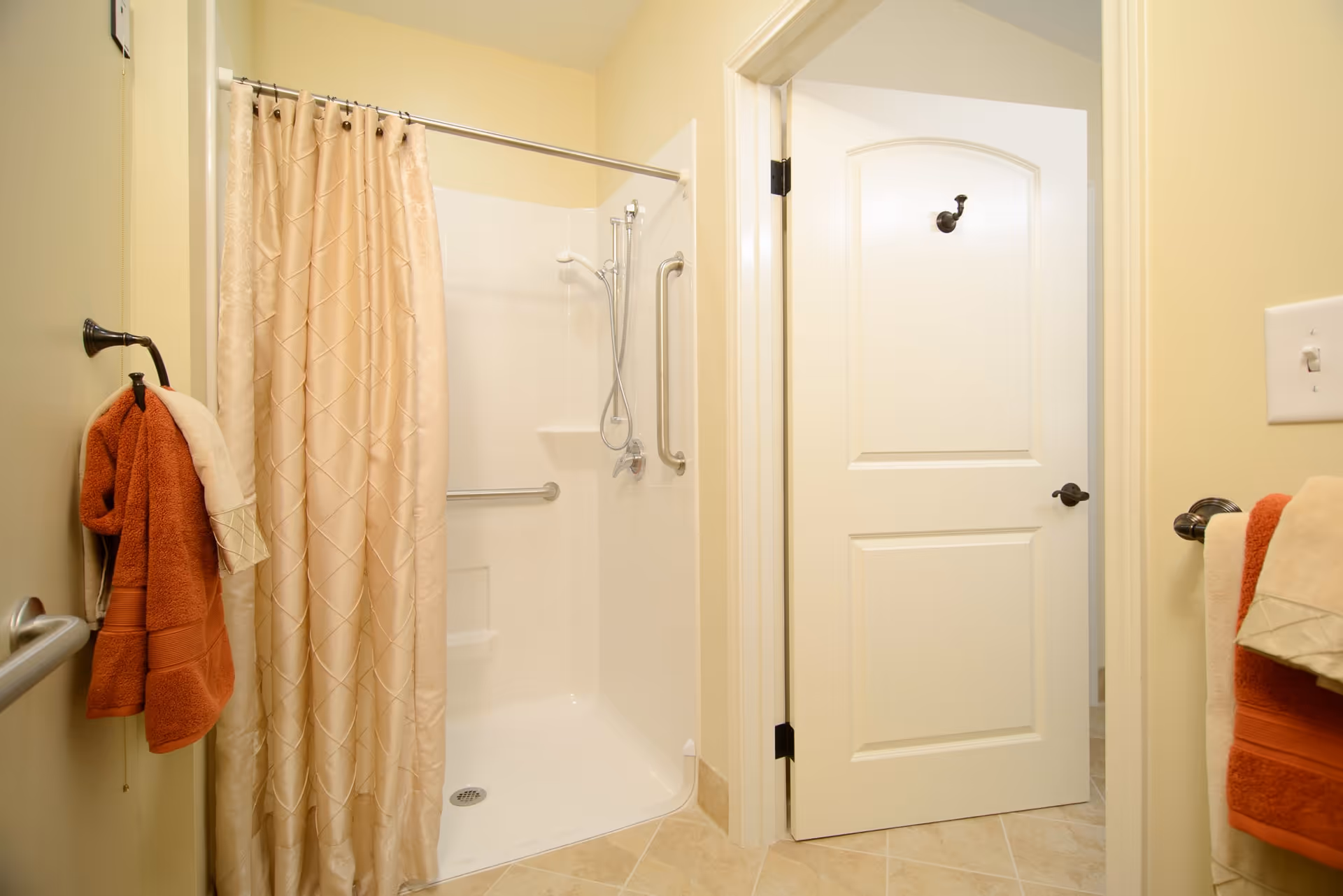 A bathroom with a walk-in shower featuring a beige patterned shower curtain, grab bars, and a handheld showerhead. There are orange and beige towels hanging on towel racks and a white door with a black hook on it. The walls are painted light yellow and the floor is tiled.