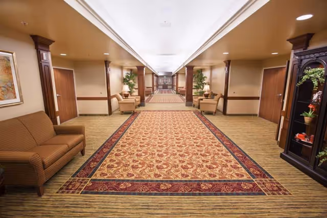 Carpeted interior hallway with a patterned runner, seating areas, columns, and decorative plants.
