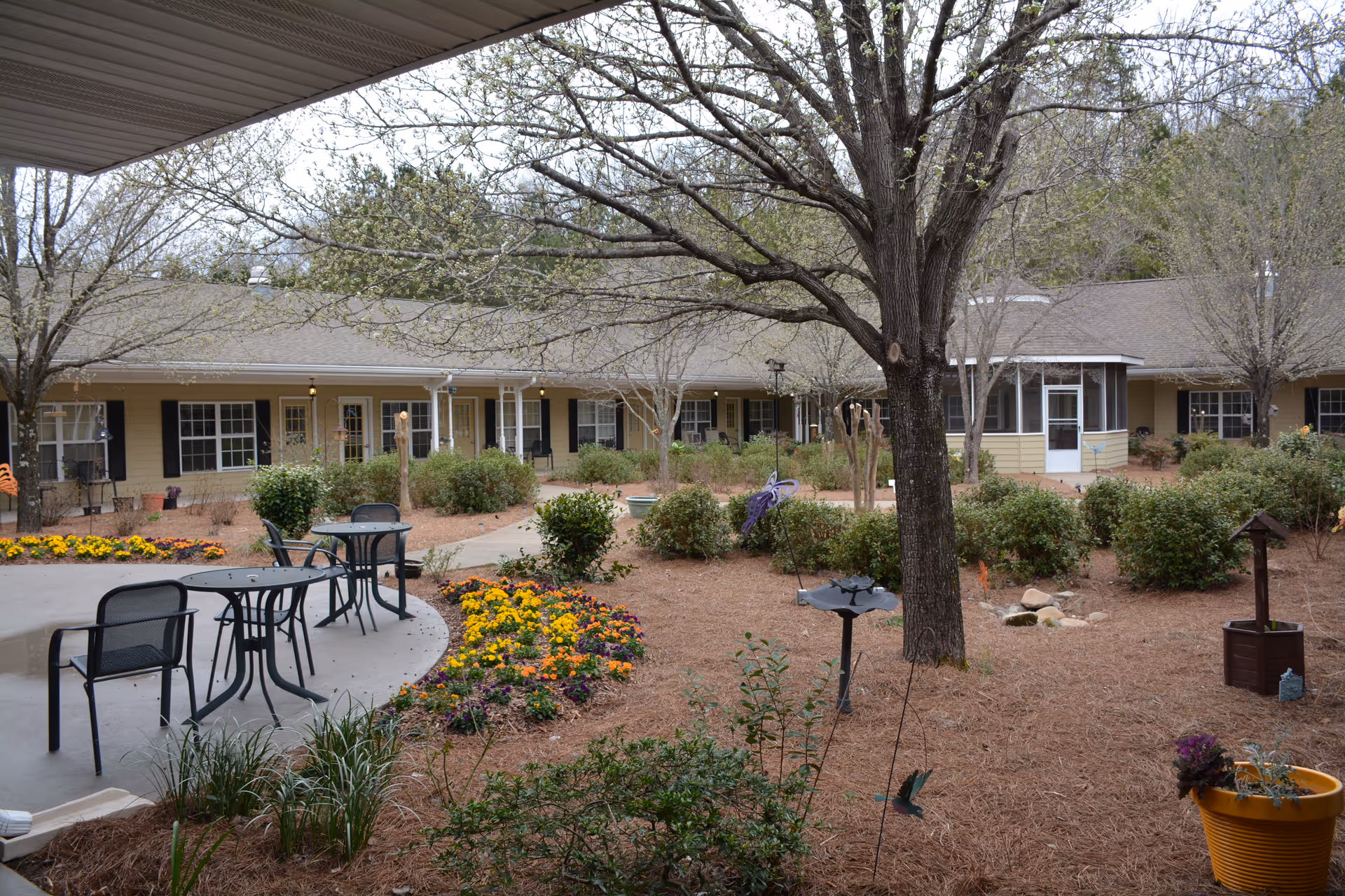 Courtyard with patio tables and chairs, flowerbeds, trees, and a surrounding single-story senior living building.