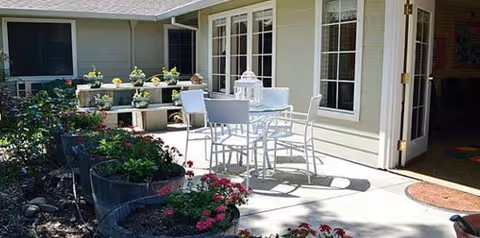 Outdoor patio area with a white metal table and four chairs on a concrete surface next to a building with large windows. There are flower beds with blooming plants and greenery surrounding the patio, and a white decorative lantern is placed on the table.