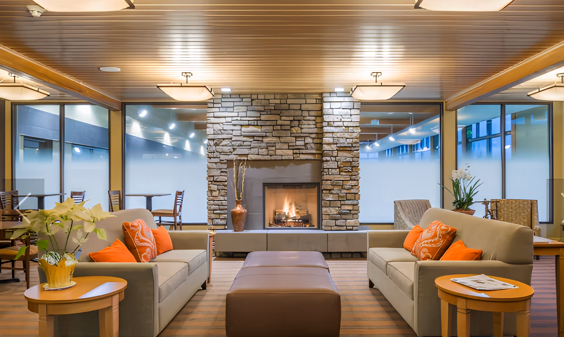 Bright communal lounge with sofas, orange pillows, a stone fireplace, and large windows.