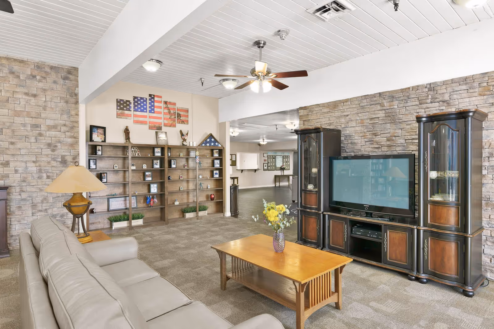 A cozy living room area in a senior living facility with a beige leather sofa, a wooden coffee table with a vase of yellow flowers, and a large dark wood entertainment center holding a flat-screen TV. The walls have a stone accent and there is a ceiling fan with lights. In the background, there is a wooden shelving unit displaying patriotic decorations and framed photos, including an American flag artwork.