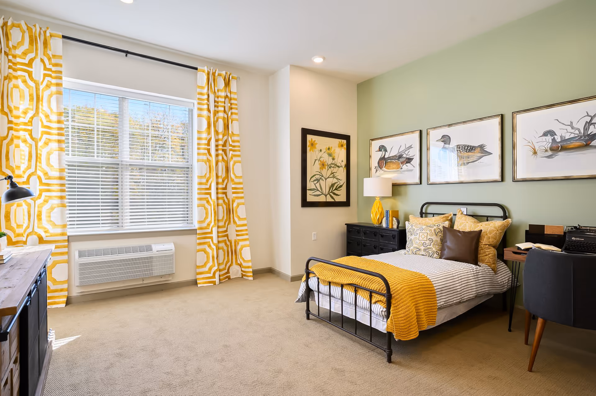 A bright and cozy bedroom with a single black metal bed frame, striped bedding, and yellow throw blanket and pillows. The room features a green accent wall with three framed bird illustrations and a floral painting on an adjacent wall. There is a black dresser with a yellow lamp and books, a wooden desk with a black chair, and large windows with yellow patterned curtains letting in natural light.