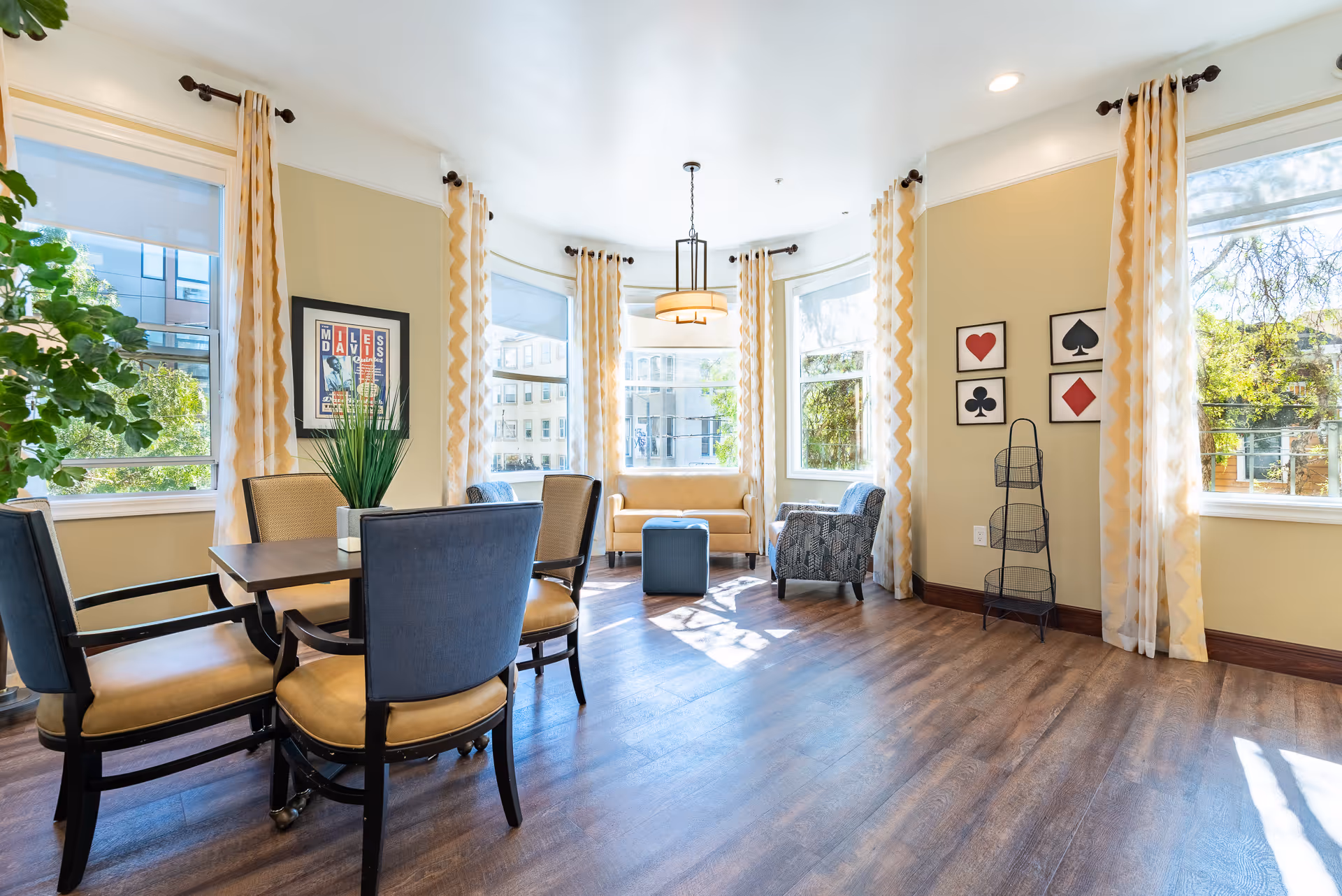 Bright and spacious common area with large windows letting in natural light, featuring a wooden table with four chairs, two armchairs, a small sofa, and a blue ottoman. The walls are decorated with framed artwork and light-colored curtains hang from dark curtain rods.