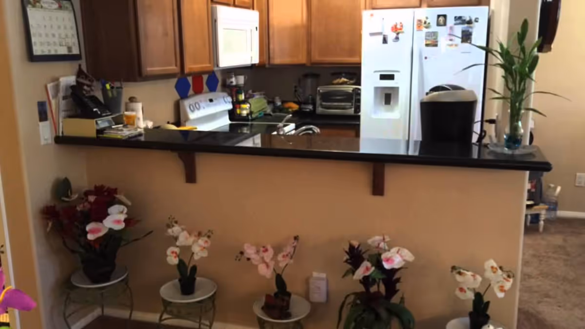 View of a kitchen area with wooden cabinets, a white refrigerator with magnets, a stove, a toaster oven, and a coffee maker on a black countertop. In front of the counter, there are five small potted orchid plants placed on small round tables against a beige wall.