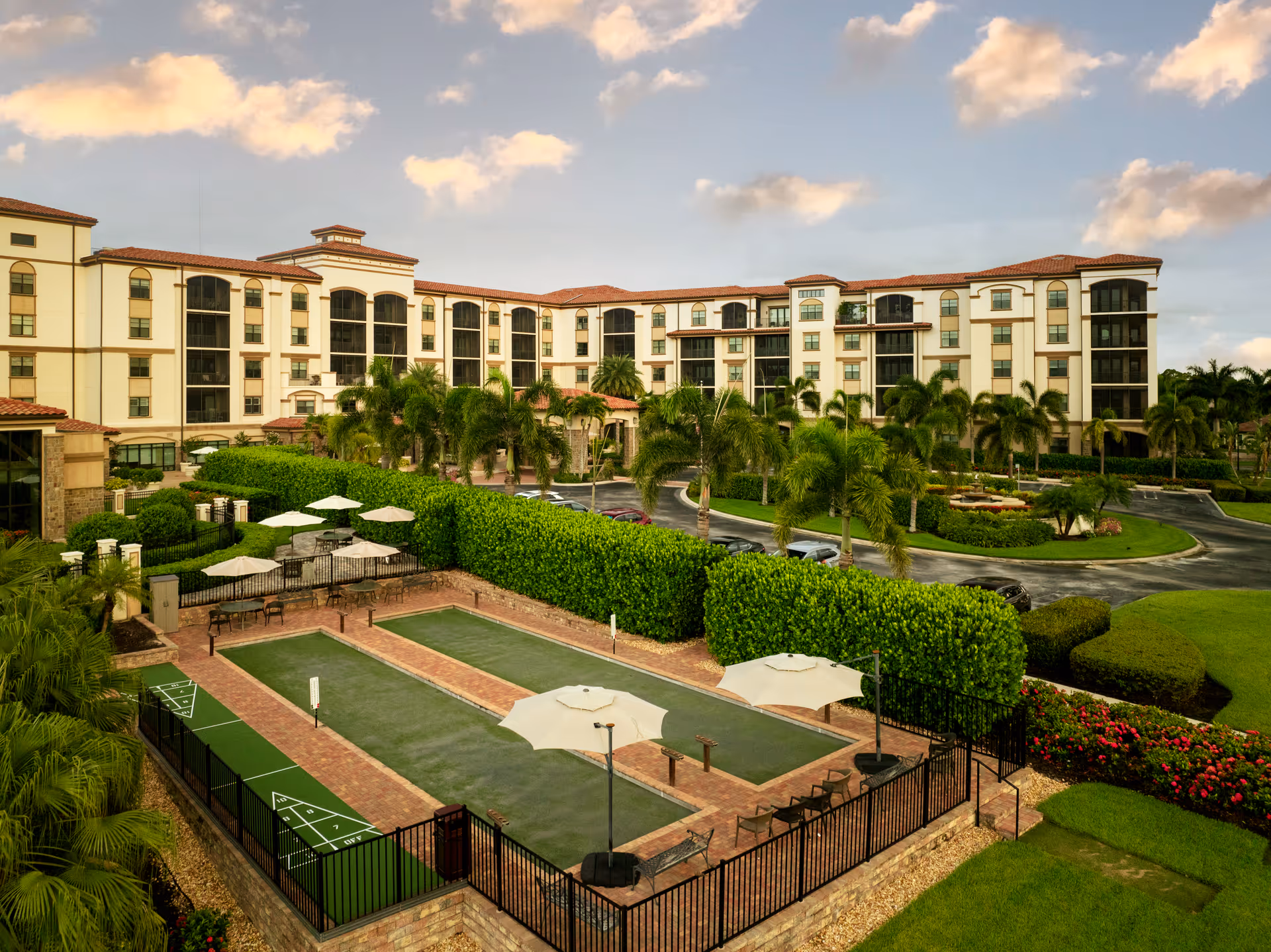 Front exterior of a Mediterranean-style senior living building with palm trees, bocce courts, umbrellas, and a circular driveway.