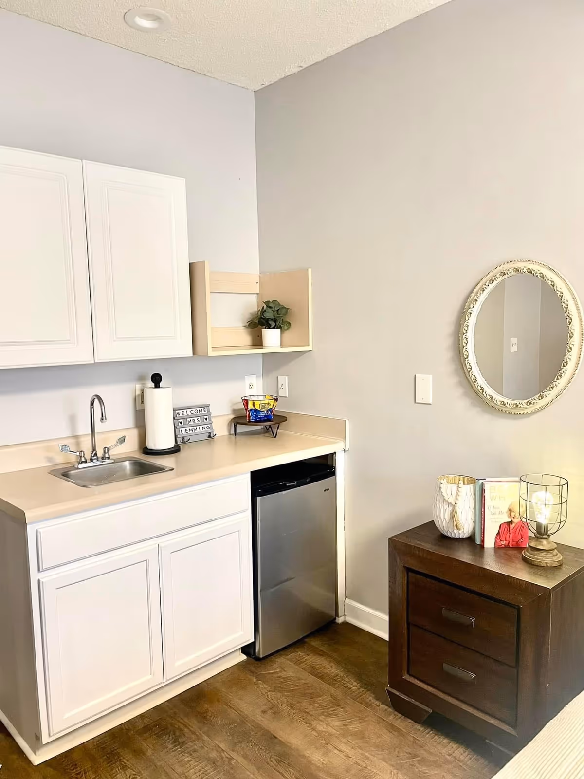 A small kitchenette area with white cabinets, a stainless steel sink, a mini refrigerator, and a countertop with a paper towel holder, a small plant, and a snack bowl. To the right, there is a wooden nightstand with two drawers, a decorative lamp, a candle holder, and a book. An oval mirror with a decorative frame hangs on the wall above the nightstand.
