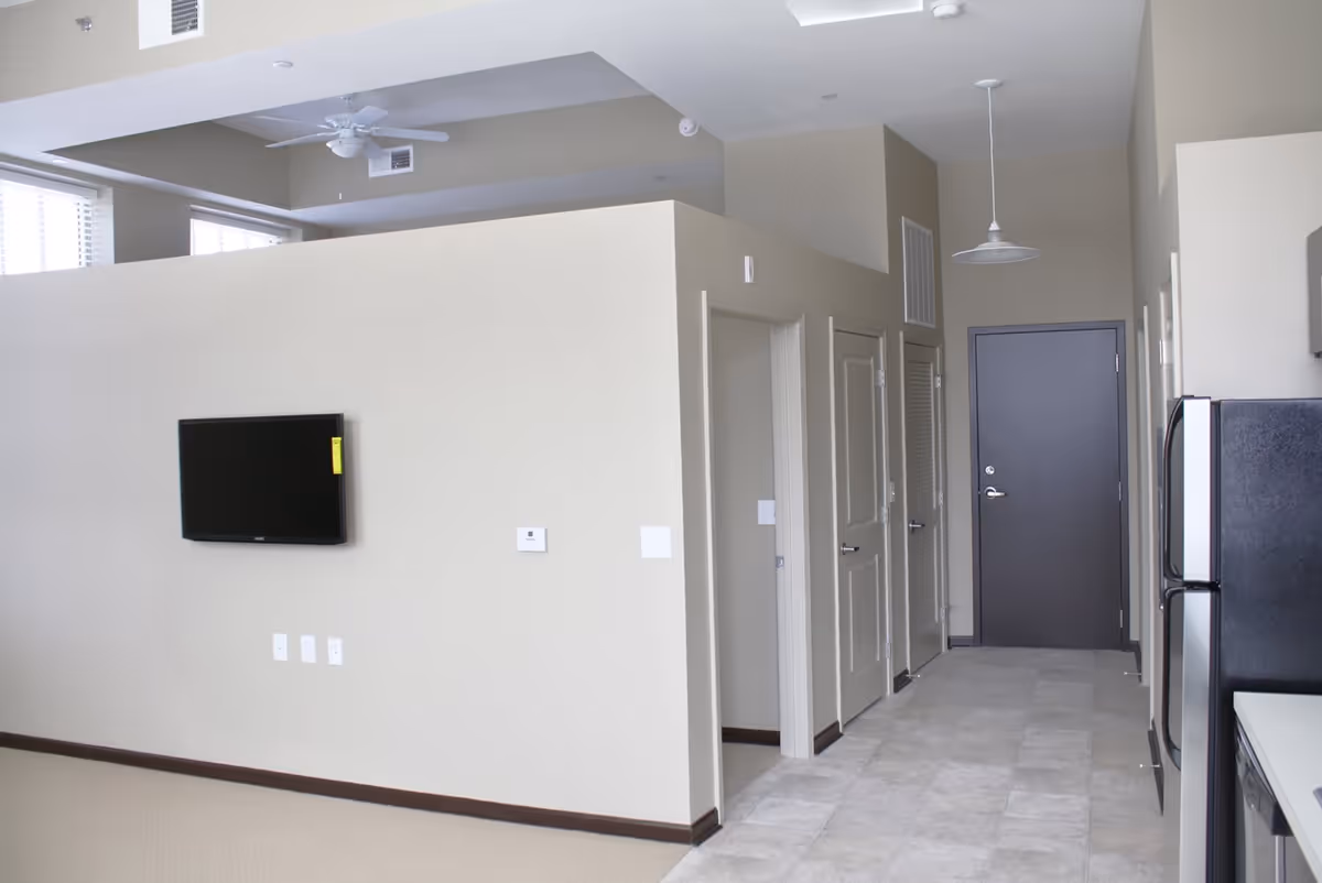 Interior view of a modern apartment or facility unit showing a hallway with multiple doors, a mounted flat-screen TV on a beige wall, a ceiling fan, and part of a kitchen area with a refrigerator and countertop visible on the right.