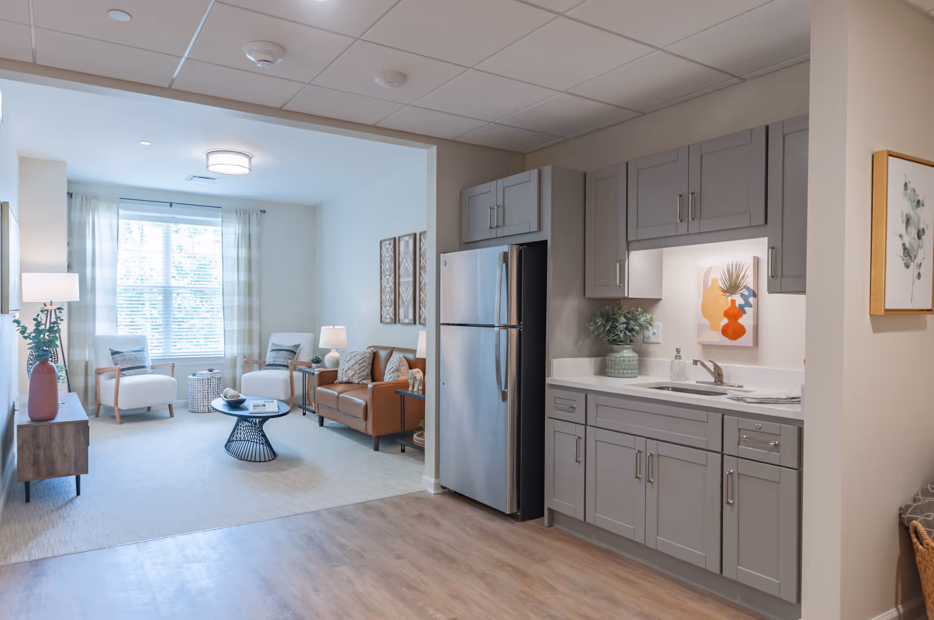 A bright and modern interior space featuring a small kitchen area with gray cabinets, a stainless steel refrigerator, and a sink. Adjacent to the kitchen is a cozy living room with a brown leather sofa, two white armchairs, a round black coffee table, and a large window with sheer curtains allowing natural light to fill the room.