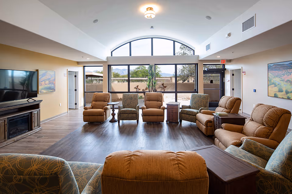 Bright communal living room with a semicircular arrangement of recliners and armchairs facing large arched windows overlooking a courtyard and distant mountains.