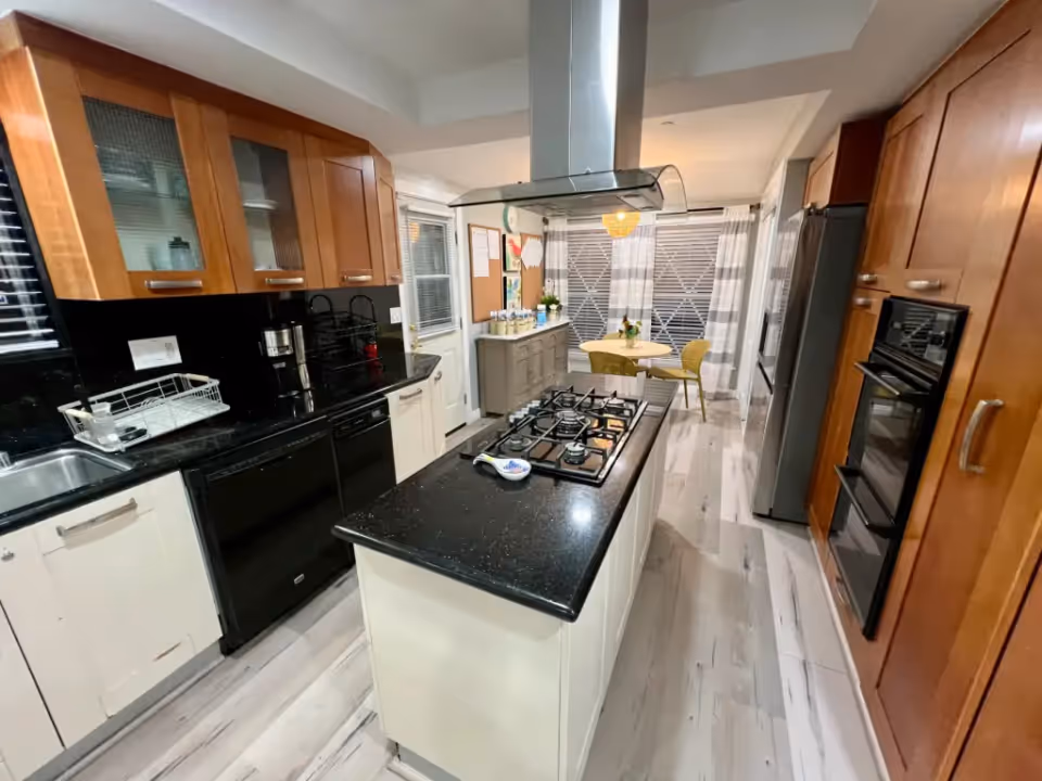 Modern kitchen with wooden upper cabinets and white lower cabinets, black countertops, a central island with a gas stove and overhead range hood, stainless steel refrigerator, built-in double oven, and a small dining area with a round table and two chairs near large windows with striped curtains.
