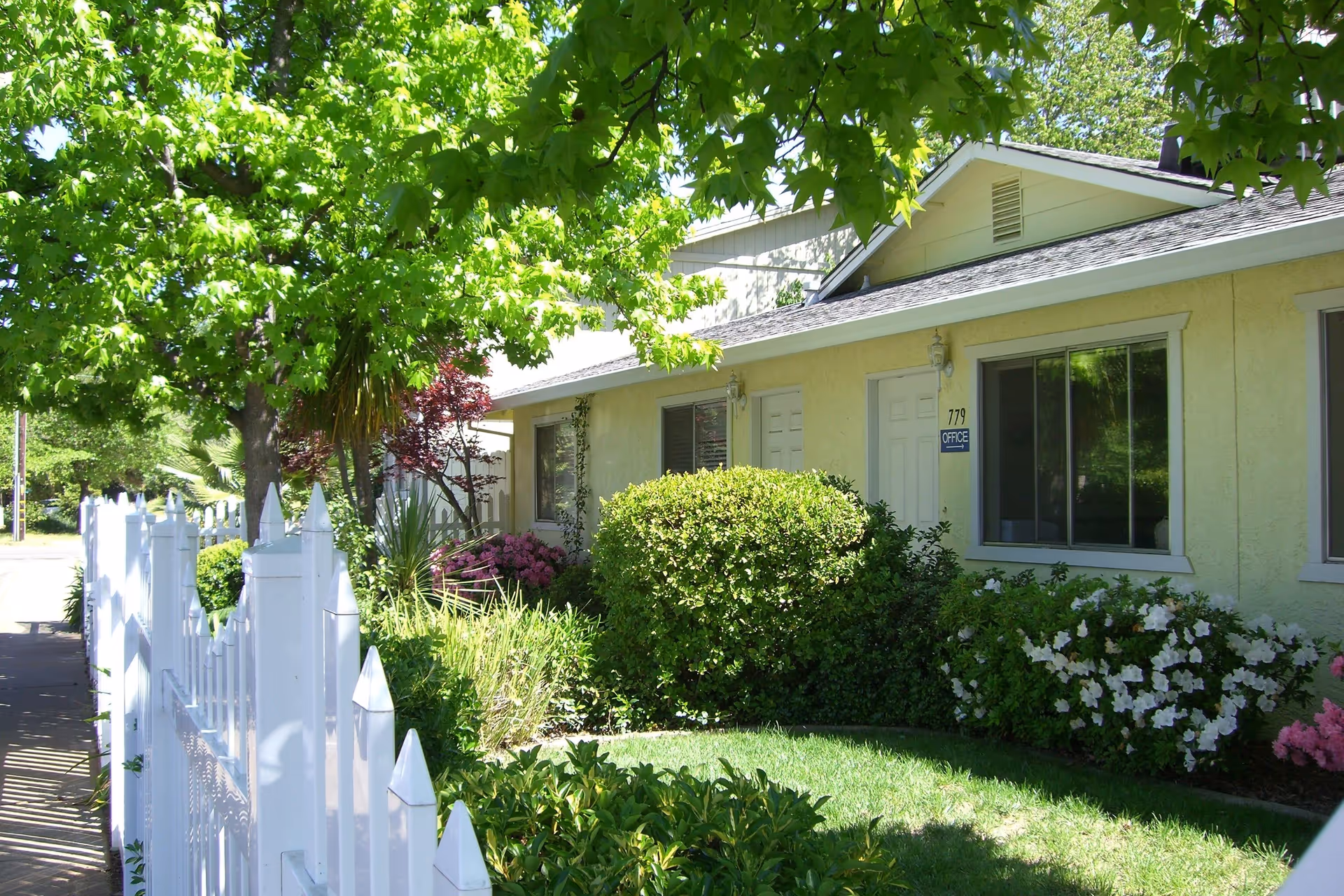Exterior view of a single-story yellow building with white trim, surrounded by green bushes, flowering plants, and trees. A white picket fence runs along the sidewalk in front of the building. A sign near the door reads '779 OFFICE'.
