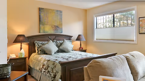 Bright bedroom featuring a wooden bed with patterned bedding, two nightstands with lamps, a window with blinds, and a couch in the foreground.