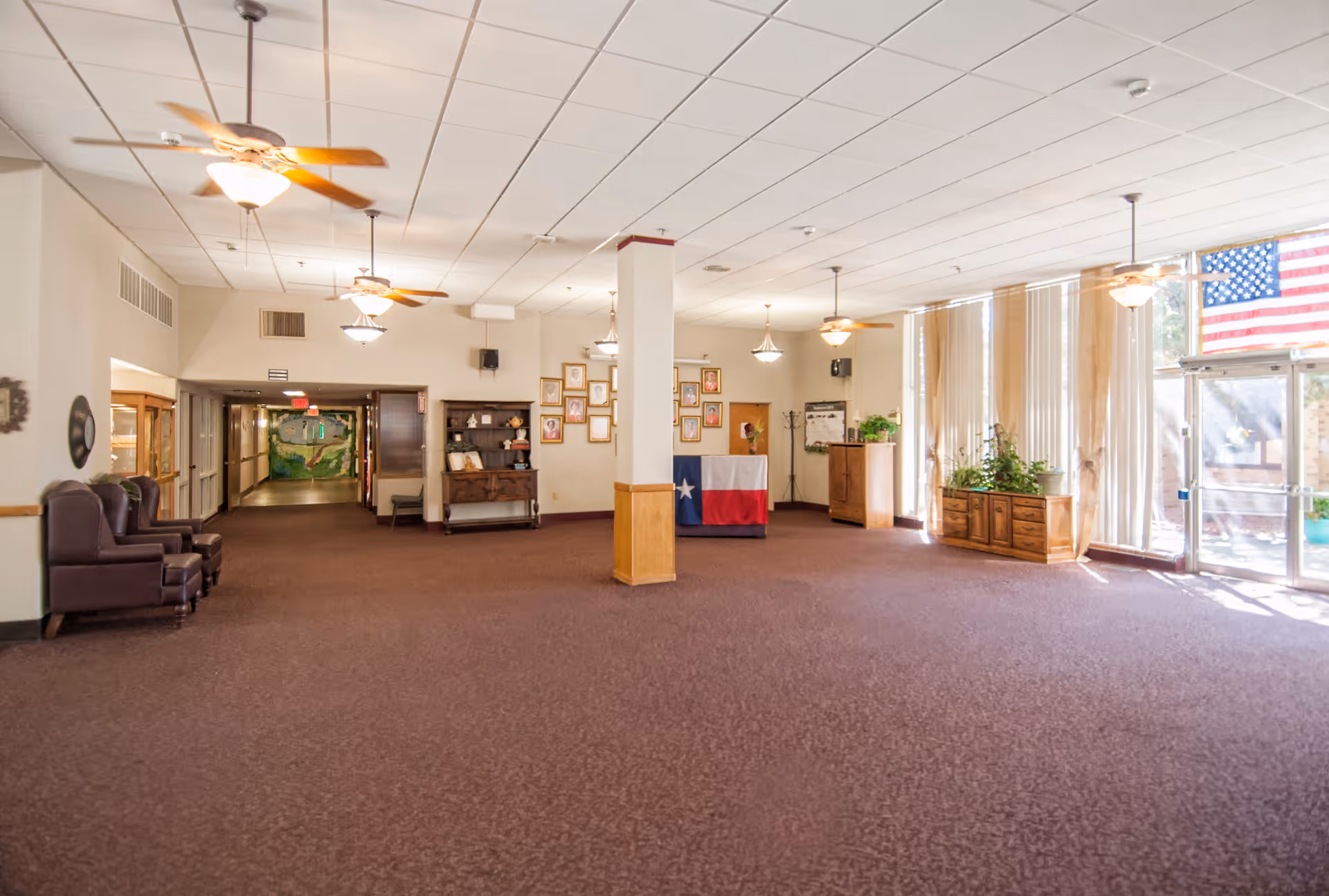 A spacious common area in a nursing home with brown carpet, beige walls, and a white ceiling with ceiling fans and light fixtures. There are two brown armchairs on the left, a hallway in the back, and a display cabinet with framed pictures on the wall. A table covered with a Texas flag is positioned near the center back wall, which is decorated with multiple framed photos. Large windows with vertical blinds and plants are on the right side, next to a glass door with an American flag hanging above it.