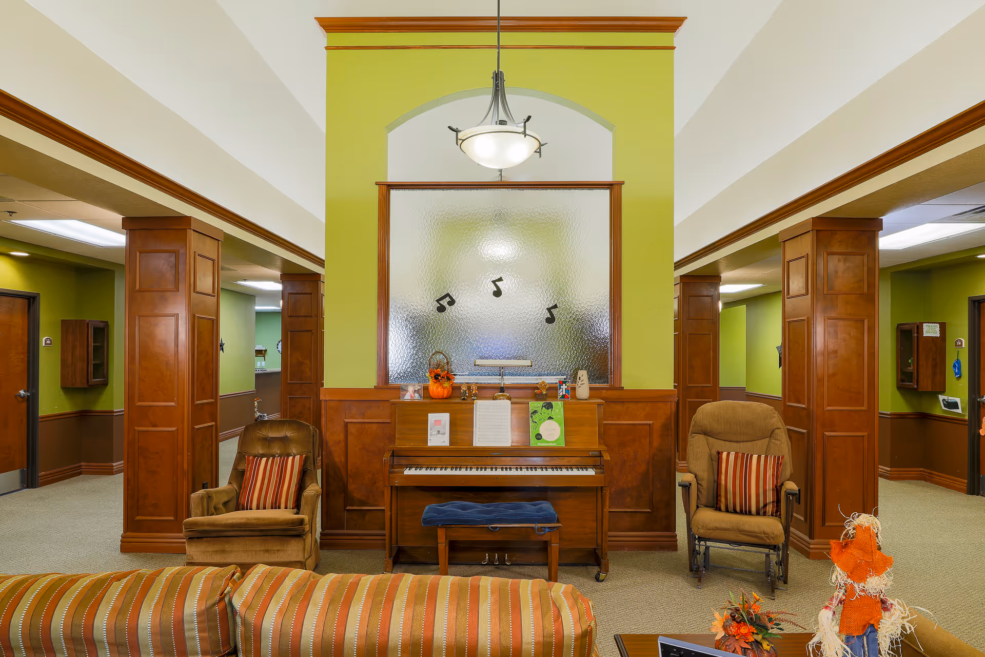 Interior common area of a senior living facility featuring a wooden piano with sheet music and decorations on top, flanked by two cushioned armchairs with striped pillows. The walls are painted green with wood paneling and columns, and there is a hanging light fixture above the piano. A striped sofa is partially visible in the foreground.