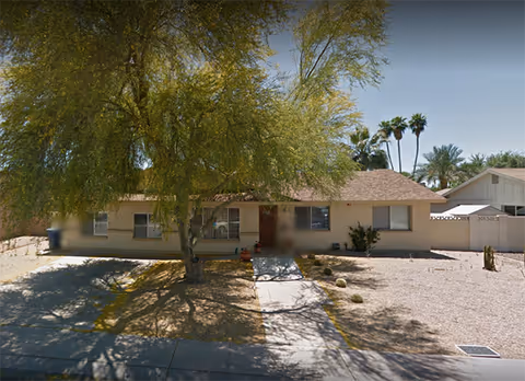 Single-story residential building with a sloped roof, surrounded by a dry landscape with gravel and a large tree in front casting shadows on the driveway and walkway leading to the entrance.