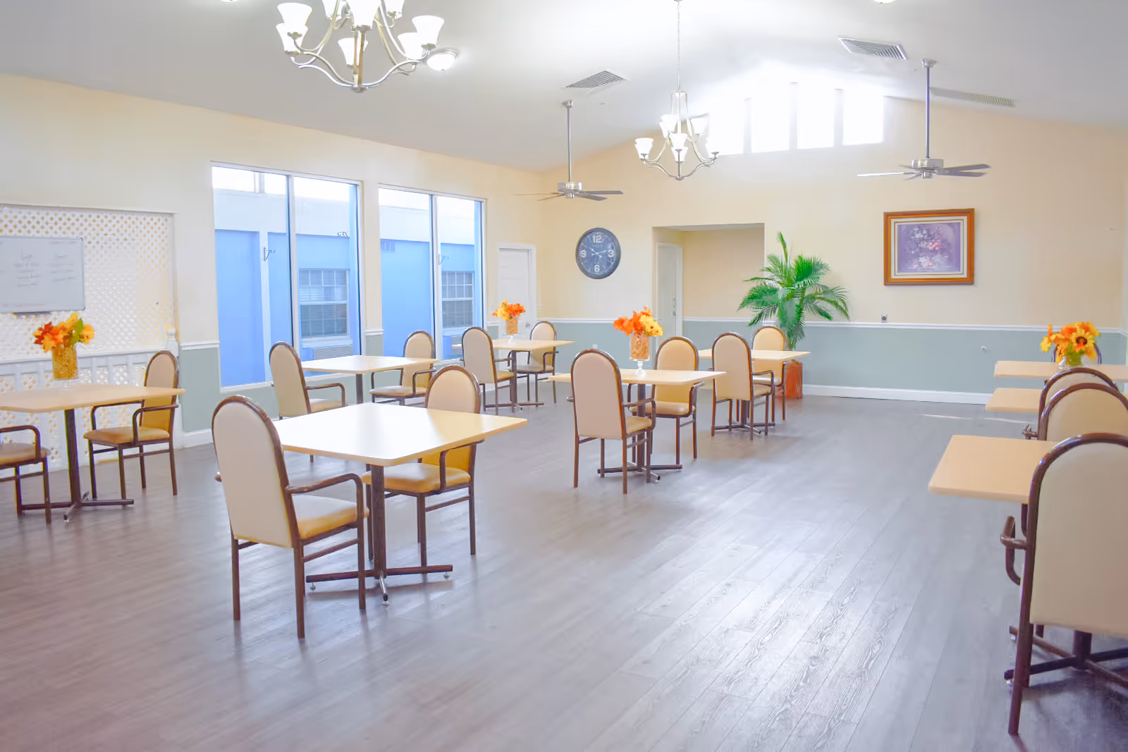 Bright spacious dining room with multiple small tables and chairs, floral centerpieces, large windows, and ceiling chandeliers.