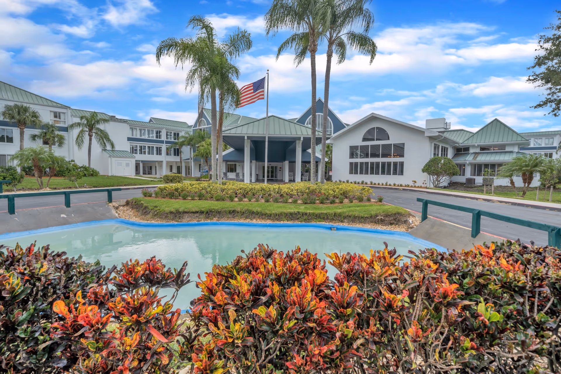 Exterior view of Discovery Village Vero Beach senior living facility with a landscaped garden, palm trees, a small pond, and an American flag in front of the building under a partly cloudy sky.