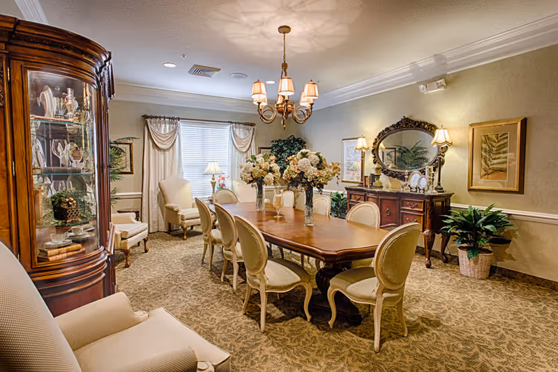 A formal dining room with a long wooden table surrounded by eight upholstered chairs. The table is decorated with three large floral arrangements. The room features a chandelier with six lampshades hanging from the ceiling, a wooden china cabinet displaying dishes and glassware, a sideboard with a large ornate mirror and two table lamps, framed artwork on the walls, and several potted plants. The room has carpeted flooring and windows with curtains allowing natural light.