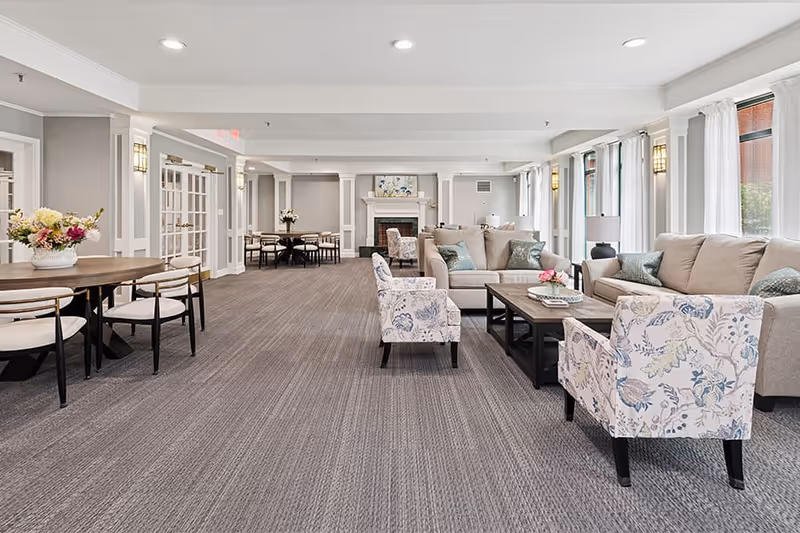 A spacious and well-lit living room area in a senior living facility featuring beige sofas, floral patterned armchairs, a wooden coffee table, and a fireplace with artwork above it. There are large windows with white curtains allowing natural light to fill the room, and additional dining tables and chairs are visible in the background.