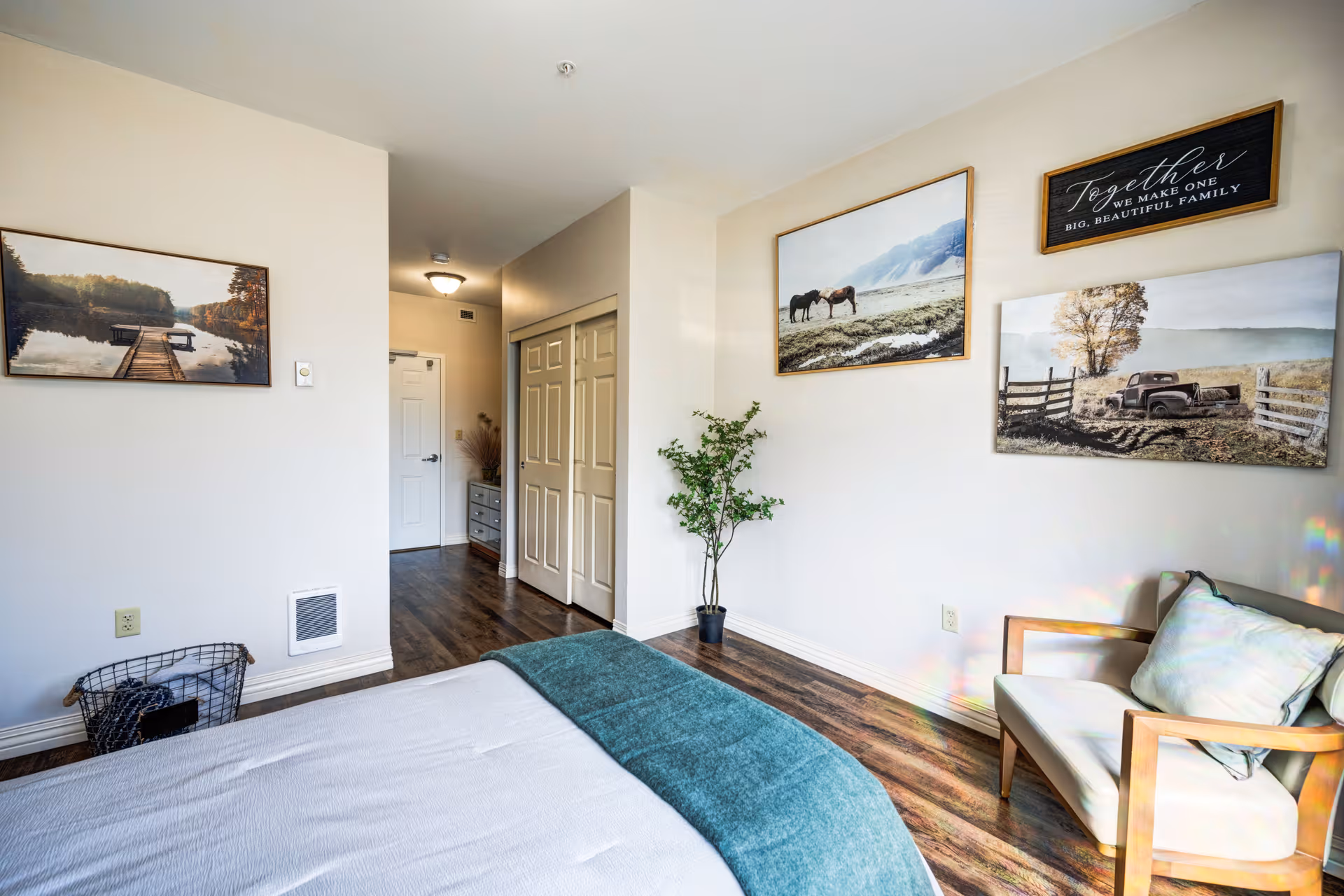A bright bedroom with a bed covered in white and teal bedding, a wooden chair with cushions, a potted plant, and several framed pictures on the walls including a sign that reads 'Together We Make One Big, Beautiful Family'. The room has wooden flooring and a hallway with a closed door and closet with sliding doors.