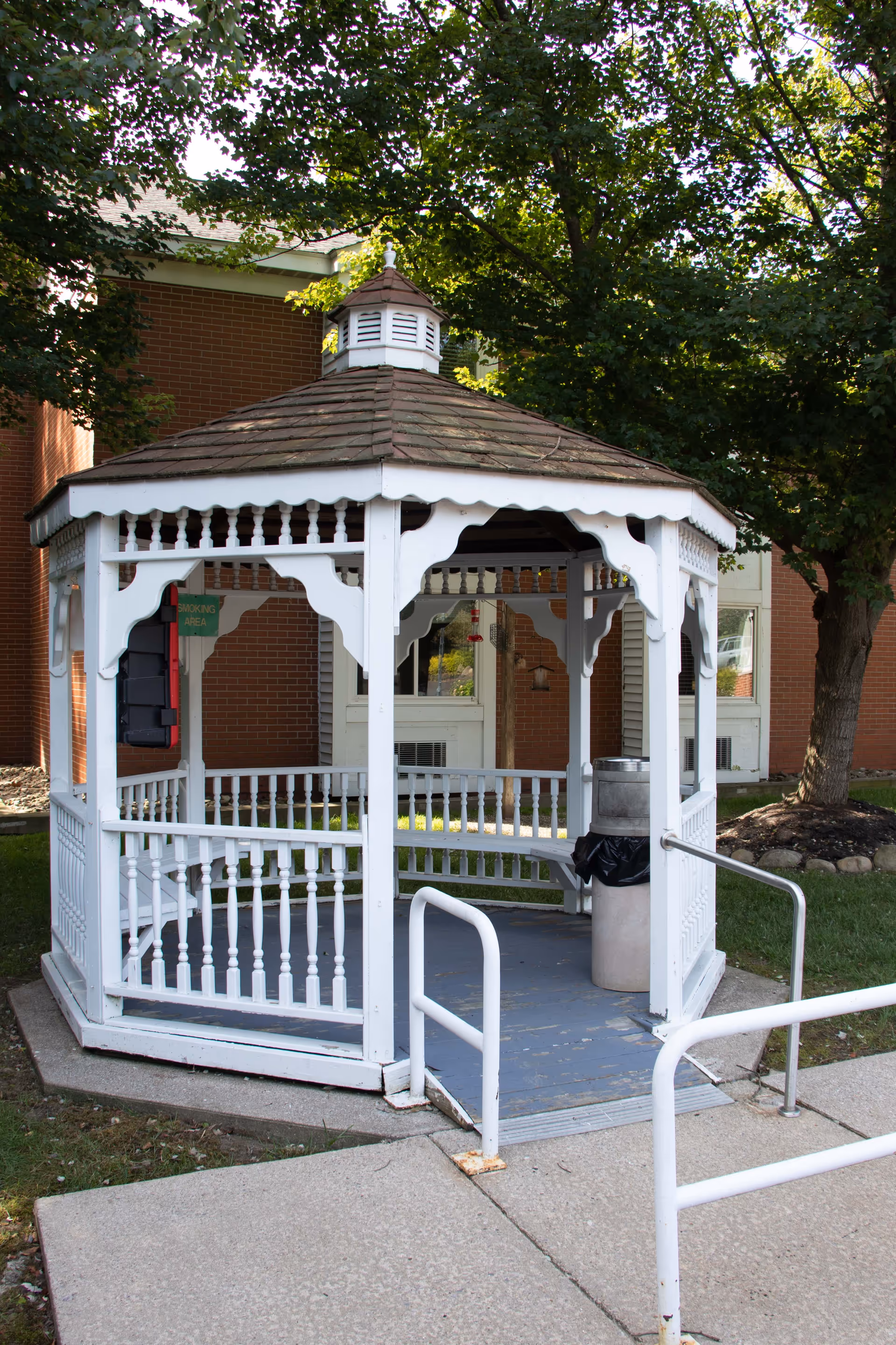 A white wooden gazebo with a shingled roof situated outdoors near a brick building. The gazebo has decorative railings and a ramp with handrails leading up to it. There is a trash can inside the gazebo and trees providing shade around it.