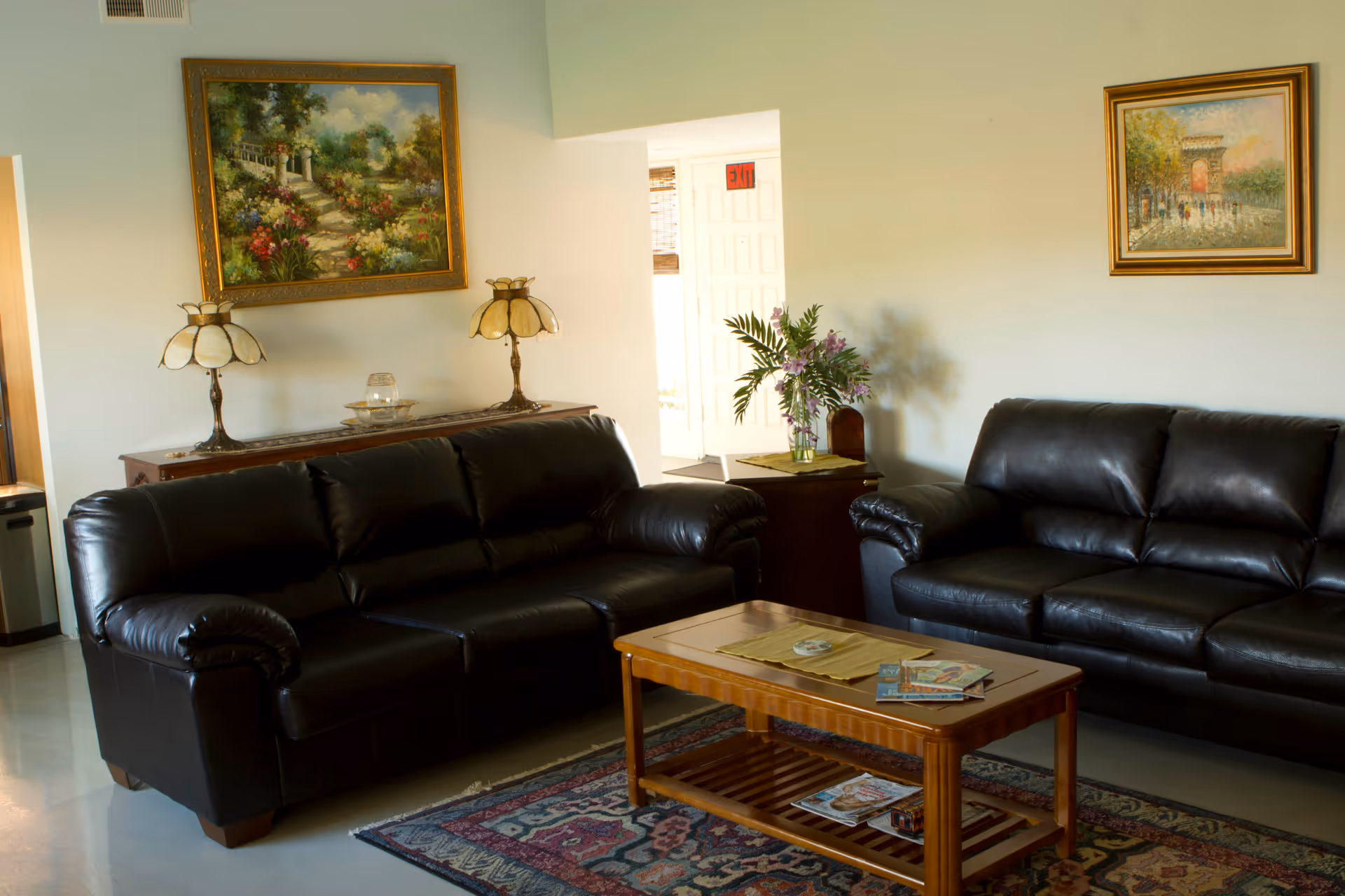 A cozy living room with two black leather sofas facing each other, a wooden coffee table with magazines and a decorative mat, a side table with a vase of flowers, two table lamps on a wooden console, and two framed paintings on the walls.