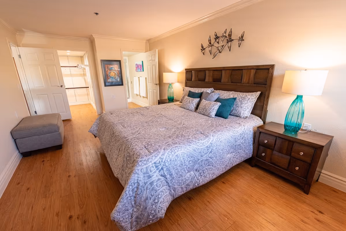 Bright furnished bedroom with a large bed, two nightstands with lamps, wood floors, and an open doorway to a closet/bathroom.