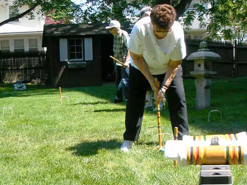 Three elderly people playing croquet on a grassy lawn in a backyard with a wooden shed and a stone lantern visible in the background.